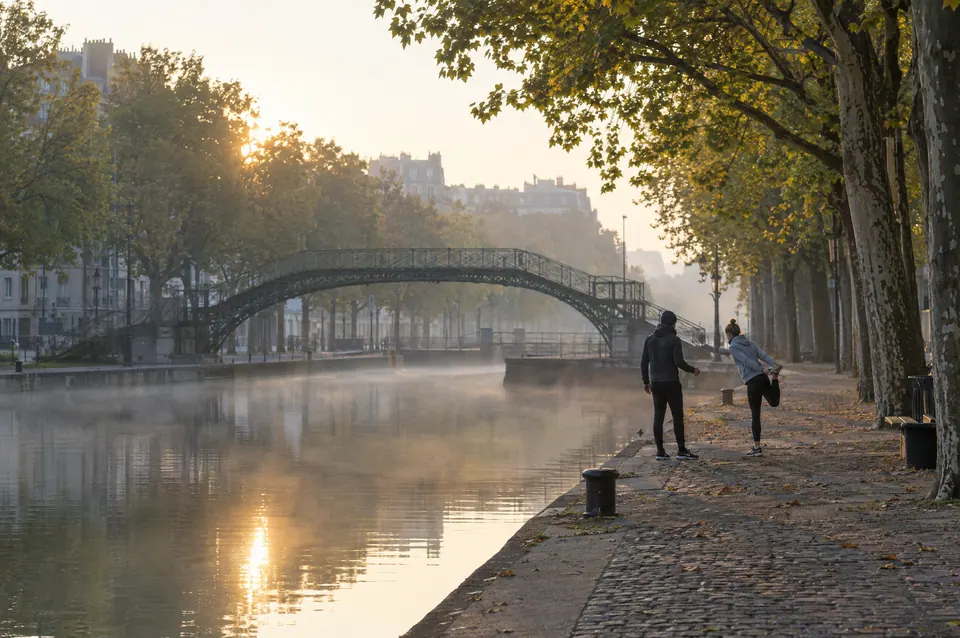 Deux personnes s’étirent sur le quai d’un canal à l’aube, sous les platanes, devant une passerelle métallique dans la brume.